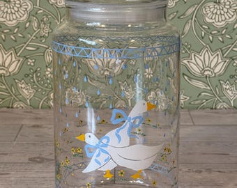 May include: A clear glass canister with a matching lid, featuring a painted design of two white geese with blue bows, yellow beaks, and feet. The jar is decorated with small yellow flowers, green grass, and blue raindrops, framed by a blue geometric border.