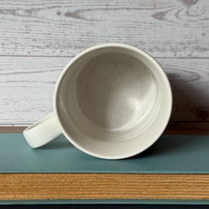 Puede incluir: Una taza de caf&eacute; de cer&aacute;mica blanca con un asa curvada. La taza est&aacute; colocada sobre un libro azul con p&aacute;ginas amarillentas y envejecidas, sobre un fondo de tablones de madera blancos.