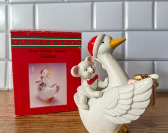 May include: A ceramic goose figurine with a mouse riding on its back, both wearing Santa hats. The goose has a yellow beak and feet. The figurine is next to a red box that reads "Hand Painted Ceramic Goose."