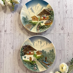 May include: Two decorative ceramic plates featuring a mountain village scene. Each plate depicts a detailed relief of wooden chalets, a road, and snow-capped mountains, with a blue and white sky background. The plates are round and have a raised edge.