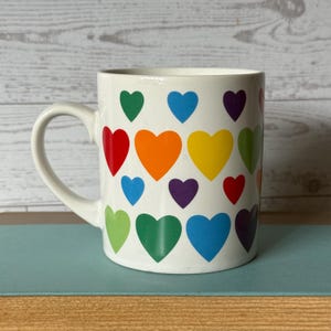 Puede incluir: Taza de cer&aacute;mica blanca decorada con corazones coloridos en un patr&oacute;n de arco&iacute;ris. Los corazones son en tonos de rojo, naranja, amarillo, verde, azul y morado. La taza tiene un asa curva.