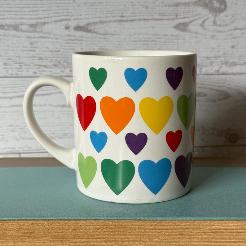 Puede incluir: Taza de cer&aacute;mica blanca decorada con corazones coloridos en un patr&oacute;n de arco&iacute;ris. Los corazones son en tonos de rojo, naranja, amarillo, verde, azul y morado. La taza tiene un asa curva.