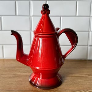 May include: A vibrant red ceramic teapot with a glossy finish. The teapot features a conical lid with a decorative finial, a curved handle, and a spout. The base and spout have a darker, shaded effect. The teapot is displayed on a wooden surface.