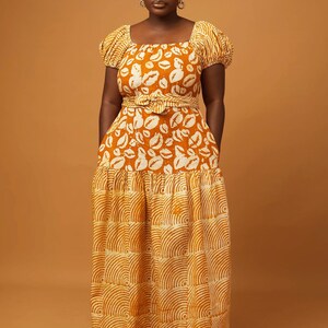 May include: A long, flowing maxi dress with a fitted bodice and a flowing skirt. The dress is made of a lightweight fabric with a printed pattern of white lips on a mustard yellow background. The skirt has a tiered design with a different pattern of white curved lines on a mustard yellow background.