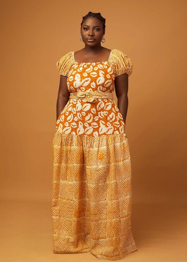 May include: A long, flowing maxi dress with a fitted bodice and a flowing skirt. The dress is made of a lightweight fabric with a printed pattern of white lips on a mustard yellow background. The skirt has a tiered design with a different pattern of white curved lines on a mustard yellow background.
