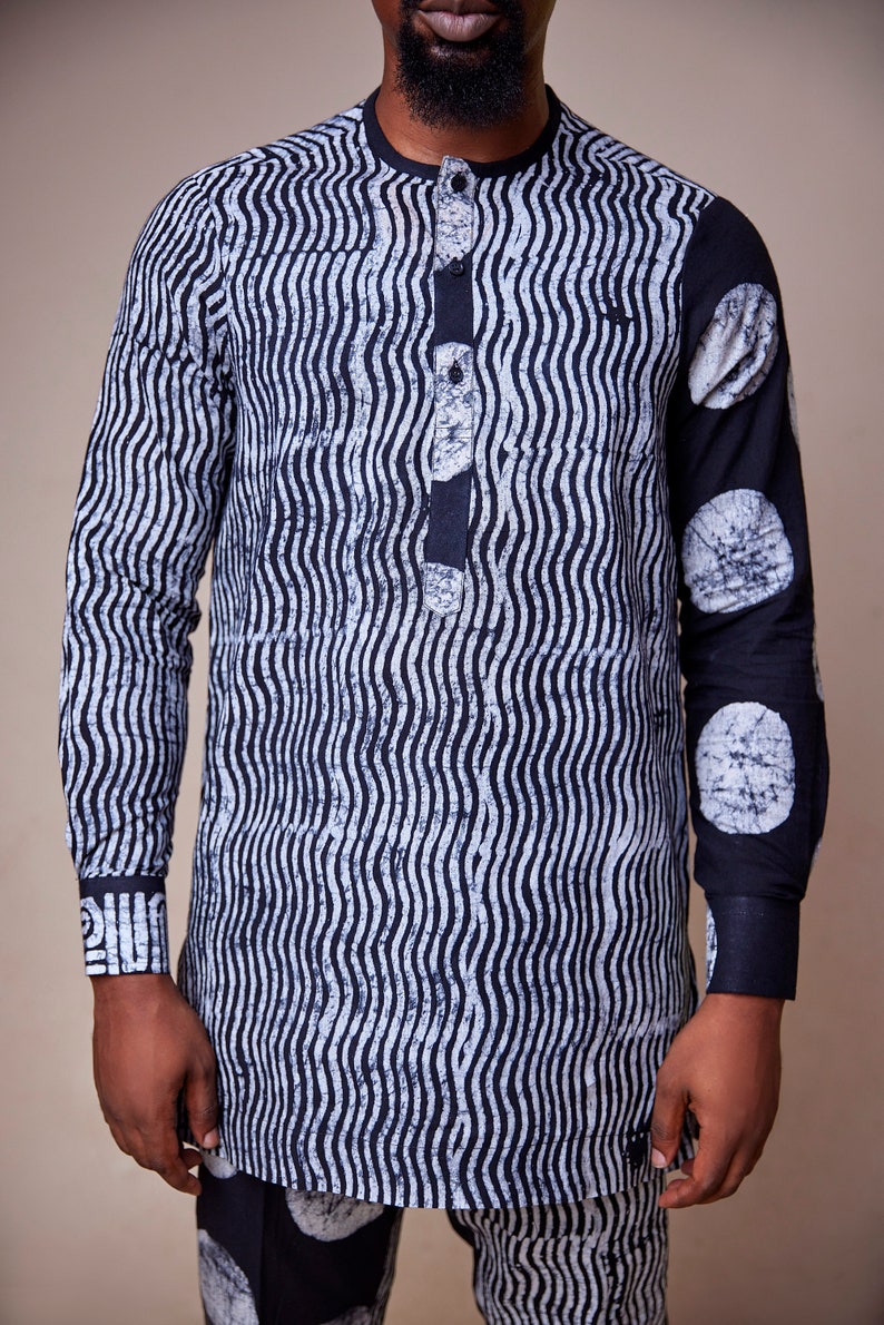 Pu&ograve; includere: Una camicia a maniche lunghe bianca e nera con un motivo ondulato e un colletto abbottonato. La camicia ha un motivo circolare sulla manica destra.