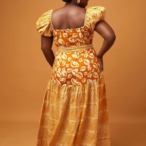May include: A long, tiered maxi dress with a fitted bodice and puff sleeves. The dress is made of a yellow and white patterned fabric with a geometric design. The dress has a square neckline and a fitted waist.