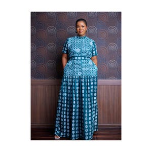 May include: A teal and white patterned maxi dress with short sleeves and a high neckline. The dress features a geometric design on the bodice and a flowing skirt with a repeating oval pattern. The dress has pockets.