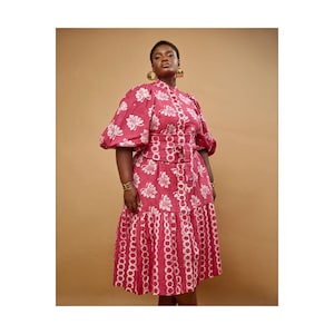 May include: A red midi dress with white floral patterns and a matching belt. The dress features a high neckline, short puff sleeves, and a tiered skirt with a unique cutout design. Gold hoop earrings and a bracelet complete the look.