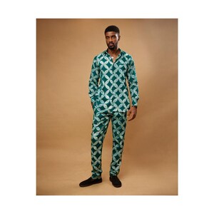 Op de afbeelding: Een bijpassende tweedelige outfit met een shirt met lange mouwen en een broek. De set heeft een wit geometrisch patroon op een teal achtergrond. De outfit wordt gedragen met zwarte schoenen.