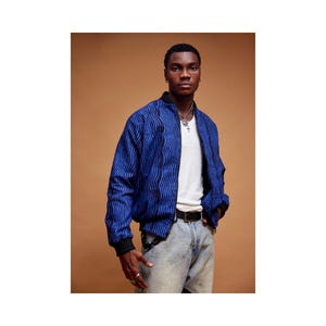 May include: A man wearing a blue and black patterned bomber jacket with a white t-shirt underneath. He is wearing blue jeans and a black belt.