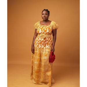 May include: A woman wearing a long yellow and white dress with a lip print pattern. The dress has a fitted bodice and a flowing skirt. She is holding a small red beaded bag.