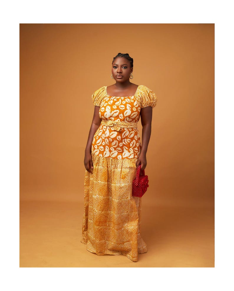 May include: A woman wearing a long yellow and white dress with a lip print pattern. The dress has a fitted bodice and a flowing skirt. She is holding a small red beaded bag.