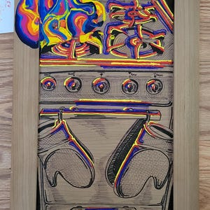 May include: A detailed illustration of a kitchen stove with oven mitts, knobs, and burners. The artwork features a vibrant, colorful flame design at the top, with a black and white stove design. The artwork is framed in a light wood.