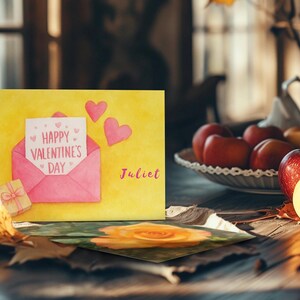 May include: A Valentine's Day card with a yellow background, featuring a pink envelope with a message that reads "Happy Valentine's Day." Two pink hearts and a small gift box are also present. The name "Juliet" is written in pink.