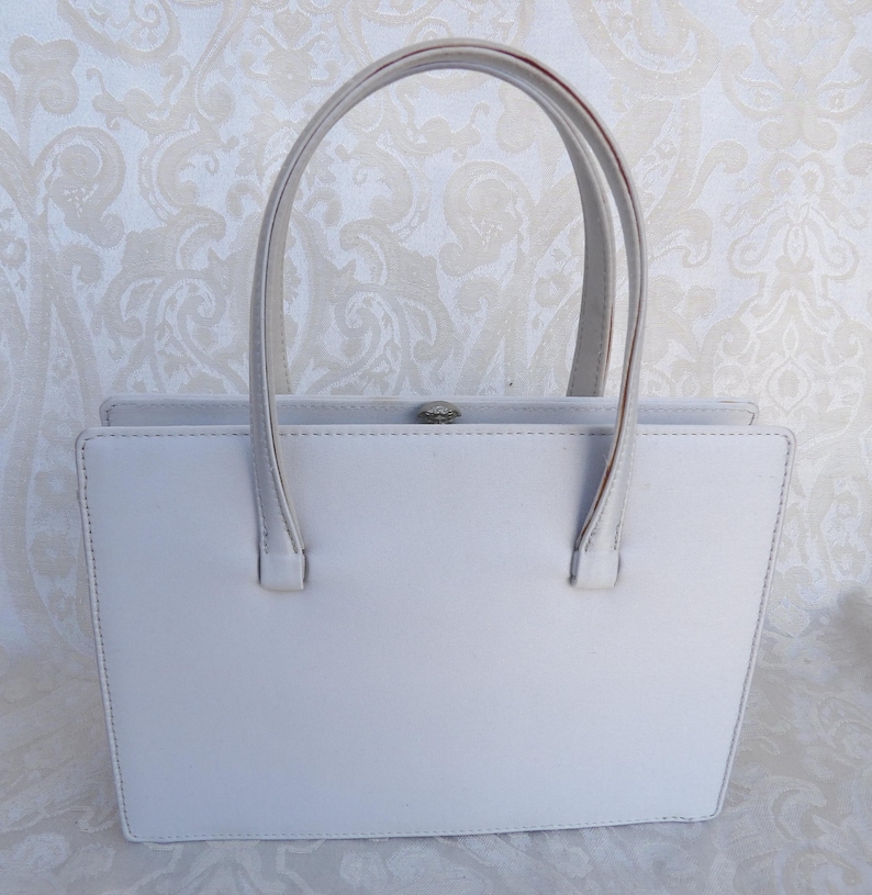 Puede incluir: Un bolso gris claro con dos asas superiores. El bolso rectangular tiene un cierre plateado y est&aacute; hecho de un material liso. Las asas est&aacute;n unidas a la parte superior de la bolsa con peque&ntilde;as fijaciones rectangulares.