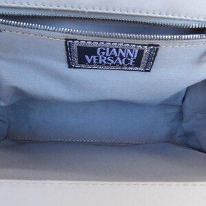 Puede incluir: Vista interior de un bolso de color claro. El interior est&aacute; forrado con una tela azul claro. Una cremallera plateada recorre la parte superior. Una etiqueta con el texto "GIANNI VERSACE" es visible dentro del bolso.