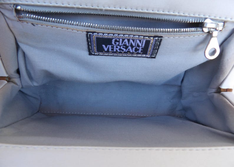 Puede incluir: Vista interior de un bolso de color claro. El interior est&aacute; forrado con una tela azul claro. Una cremallera plateada recorre la parte superior. Una etiqueta con el texto "GIANNI VERSACE" es visible dentro del bolso.