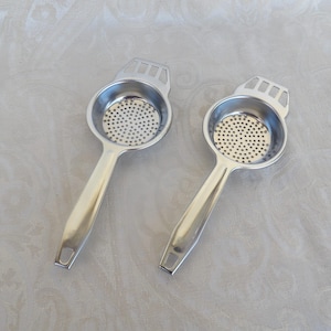 May include: Two silver-colored stainless steel tea strainers with circular perforated bowls and flat handles. Each strainer features a small rectangular cutout at the end of the handle and a decorative slotted design at the top of the bowl.
