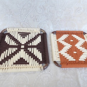 May include: Two square woven coasters. One has a brown and cream geometric pattern, the other an orange and cream design. Both have a dark brown frame. They are placed on a white patterned surface. Each coaster measures approximately 15 cm x 15 cm.
