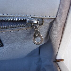 Puede incluir: Primer plano del interior de un bolso gris claro con una cremallera plateada y un tirador de cremallera met&aacute;lico. El tirador de la cremallera est&aacute; marcado con la palabra "riri". Una etiqueta que dice "MADE IN ITALY" es visible.