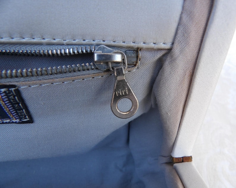 Puede incluir: Primer plano del interior de un bolso gris claro con una cremallera plateada y un tirador de cremallera met&aacute;lico. El tirador de la cremallera est&aacute; marcado con la palabra "riri". Una etiqueta que dice "MADE IN ITALY" es visible.
