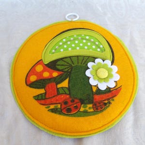 Peut inclure: Un dessous-de-plat rond en feutre avec un motif de champignons. Le dessous-de-plat a un fond jaune et une bordure verte. Les champignons sont rouges et verts avec des pois blancs. Une fleur blanche et jaune est également présente.