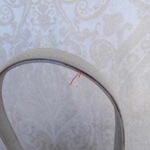 Puede incluir: Primer plano de un asa de bolso de color crema. El asa es curva y parece estar hecha de cuero o un material similar. Las costuras son visibles a lo largo de los bordes y el fondo es un papel pintado con dibujos.