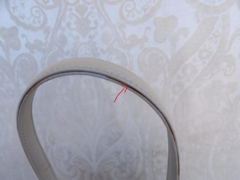 Puede incluir: Primer plano de un asa de bolso de color crema. El asa es curva y parece estar hecha de cuero o un material similar. Las costuras son visibles a lo largo de los bordes y el fondo es un papel pintado con dibujos.