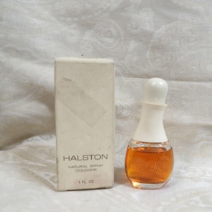 May include: A Halston natural spray cologne bottle with a white cap and amber liquid, standing next to its rectangular white box. The box features the text HALSTON, NATURAL SPRAY, COLOGNE, and 1 FL. OZ. printed in black on the front surface.