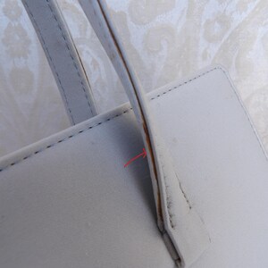 Puede incluir: Primer plano de un bolso de mano de cuero gris claro. El bolso tiene forma rectangular con un asa y costuras visibles. El asa est&aacute; sujeta al bolso con un acento marr&oacute;n. El fondo es una tela clara estampada.