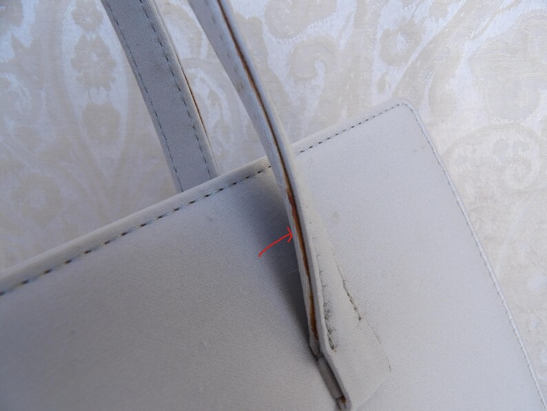 Puede incluir: Primer plano de un bolso de mano de cuero gris claro. El bolso tiene forma rectangular con un asa y costuras visibles. El asa est&aacute; sujeta al bolso con un acento marr&oacute;n. El fondo es una tela clara estampada.