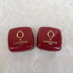 Puede incluir: Dos polveras cuadradas de color burdeos con letras doradas y un emblema dorado. El texto en las polveras dice "de LANCÔME PARIS". Las polveras están colocadas sobre una superficie blanca estampada.