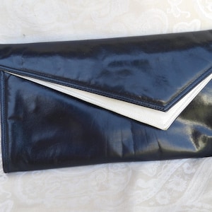 May include: A black leather clutch with a white interior flap. The clutch has a rectangular shape with a triangular flap closure. The leather appears shiny and smooth, and the overall design is simple and elegant.