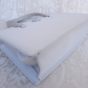 Puede incluir: Un bolso de mano rectangular blanco con un asa superior y un cierre plateado. El bolso tiene un dise&ntilde;o estructurado con costuras visibles y un acabado brillante. El bolso probablemente est&aacute; hecho de cuero o un material similar.