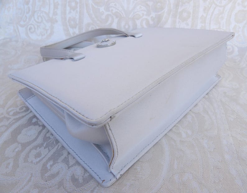 Puede incluir: Un bolso de mano rectangular blanco con un asa superior y un cierre plateado. El bolso tiene un dise&ntilde;o estructurado con costuras visibles y un acabado brillante. El bolso probablemente est&aacute; hecho de cuero o un material similar.