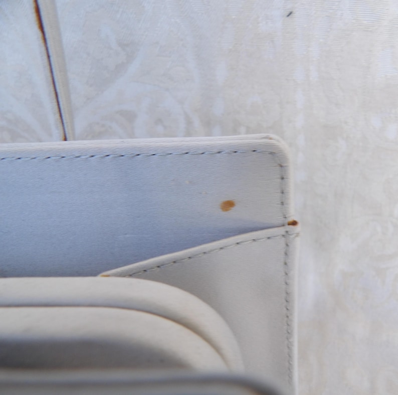 Puede incluir: Primer plano de una caja de joyer&iacute;a rectangular gris claro con un interior curvo y acolchado. La caja tiene un borde cosido y una peque&ntilde;a mancha marr&oacute;n en la superficie. El fondo es una tela blanca texturizada.