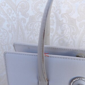 Puede incluir: Primer plano de un bolso de mano blanco con asa. El bolso tiene forma rectangular con un cierre plateado y un elemento decorativo. El asa es curva y est&aacute; sujeta a la parte superior del bolso.
