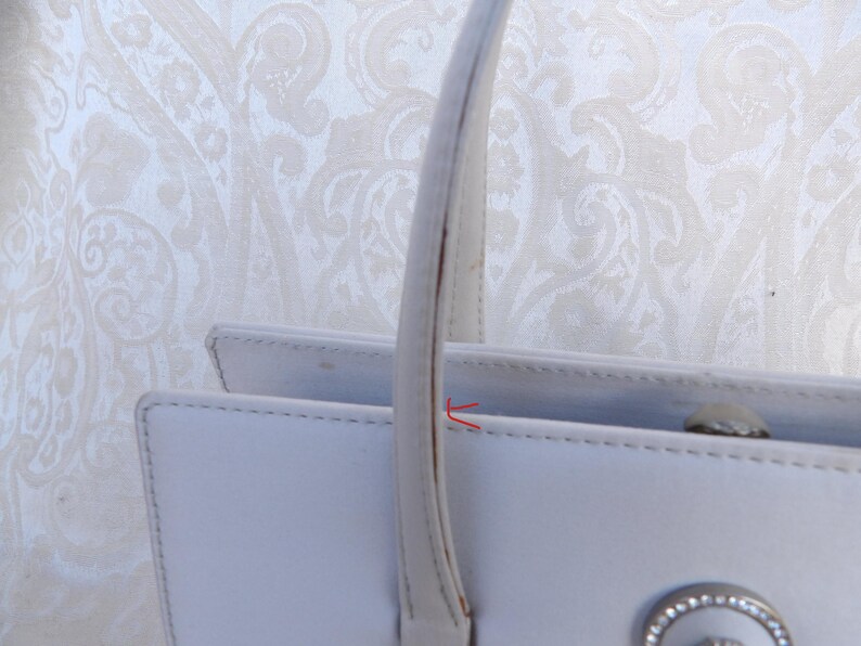 Puede incluir: Primer plano de un bolso de mano blanco con asa. El bolso tiene forma rectangular con un cierre plateado y un elemento decorativo. El asa es curva y est&aacute; sujeta a la parte superior del bolso.