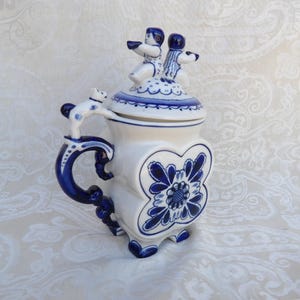 May include: A white ceramic lidded tankard featuring blue floral patterns and a sculpted handle with a small animal figure. The lid is topped with two small human figures, and the body has a four-lobed shape with blue painted details.