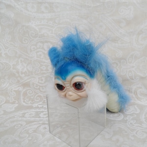 May include: A plush creature with a pale, smooth face, large dark eyes, and a small nose. It features bright blue faux fur on its head and back, with white fur accents around its cheeks. The creature is positioned on a clear acrylic stand.