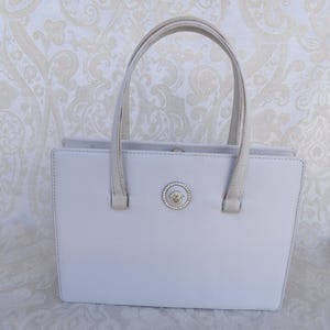 Puede incluir: Un bolso de mano gris claro con forma rectangular y dos asas. El bolso presenta un emblema plateado con un dise&ntilde;o circular y un borde decorativo. Las asas y los ribetes son de un tono gris m&aacute;s claro.