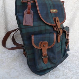 May include: A green and navy plaid backpack with brown leather trim and accents. The backpack features a flap closure, two front pockets, and a leather luggage tag. The straps and trim are brown, and the bag has a drawstring closure.