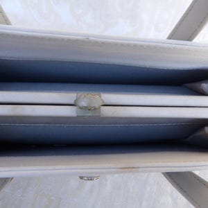 Puede incluir: Vista abierta de un bolso gris claro con un cierre plateado. El interior es de un azul-gris contrastante. El bolso tiene dos asas y una forma rectangular. El cierre es un dise&ntilde;o decorativo en forma de flor.