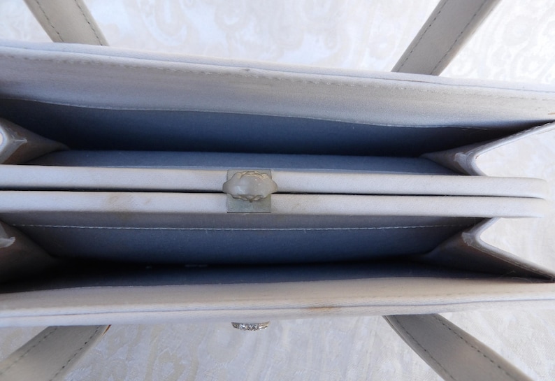 Puede incluir: Vista abierta de un bolso gris claro con un cierre plateado. El interior es de un azul-gris contrastante. El bolso tiene dos asas y una forma rectangular. El cierre es un dise&ntilde;o decorativo en forma de flor.