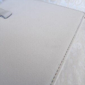 Puede incluir: Un portafolio de cuero gris claro con asa. El portafolio tiene un borde cosido y una superficie lisa, ligeramente texturizada. El asa es de color gris claro a juego.