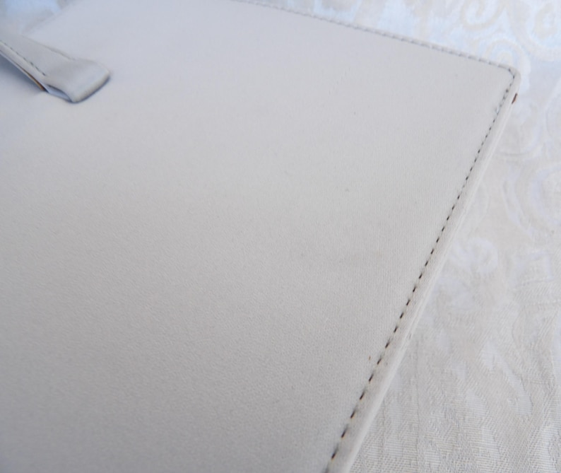 Puede incluir: Un portafolio de cuero gris claro con asa. El portafolio tiene un borde cosido y una superficie lisa, ligeramente texturizada. El asa es de color gris claro a juego.