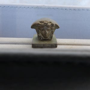 Puede incluir: Primer plano de un emblema met&aacute;lico de cabeza de Medusa sobre una base rectangular. El emblema es de color bronce claro y est&aacute; sobre un fondo azul claro y beige.