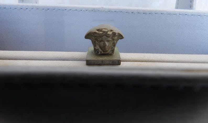 Puede incluir: Primer plano de un emblema met&aacute;lico de cabeza de Medusa sobre una base rectangular. El emblema es de color bronce claro y est&aacute; sobre un fondo azul claro y beige.