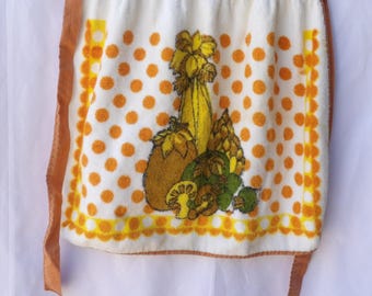Könnte beinhalten: Eine Vintage-Küchenschürze mit weißem Hintergrund und orangefarbenen Punkten. Die Schürze zeigt eine farbenfrohe Illustration verschiedener Gemüsesorten, darunter Mais, Kürbis und Pilze. Die Schürze hat braune Bindebänder und einen braunen Besatz.